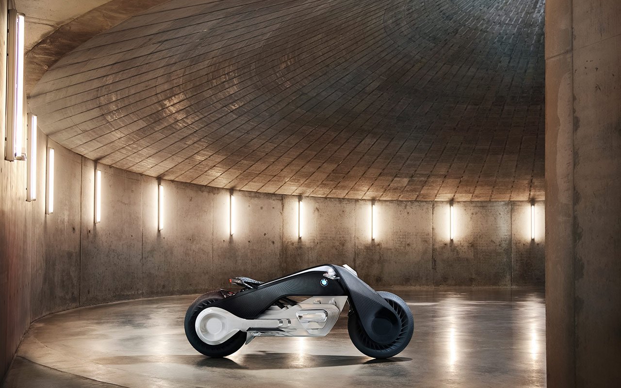 The futuristic BMW Motorrad VISION NEXT 100 concept motorcycle on display in a modern, minimalist space with a concrete floor, walls, and ceiling. Light fixtures illuminate the sleek design of the bike.